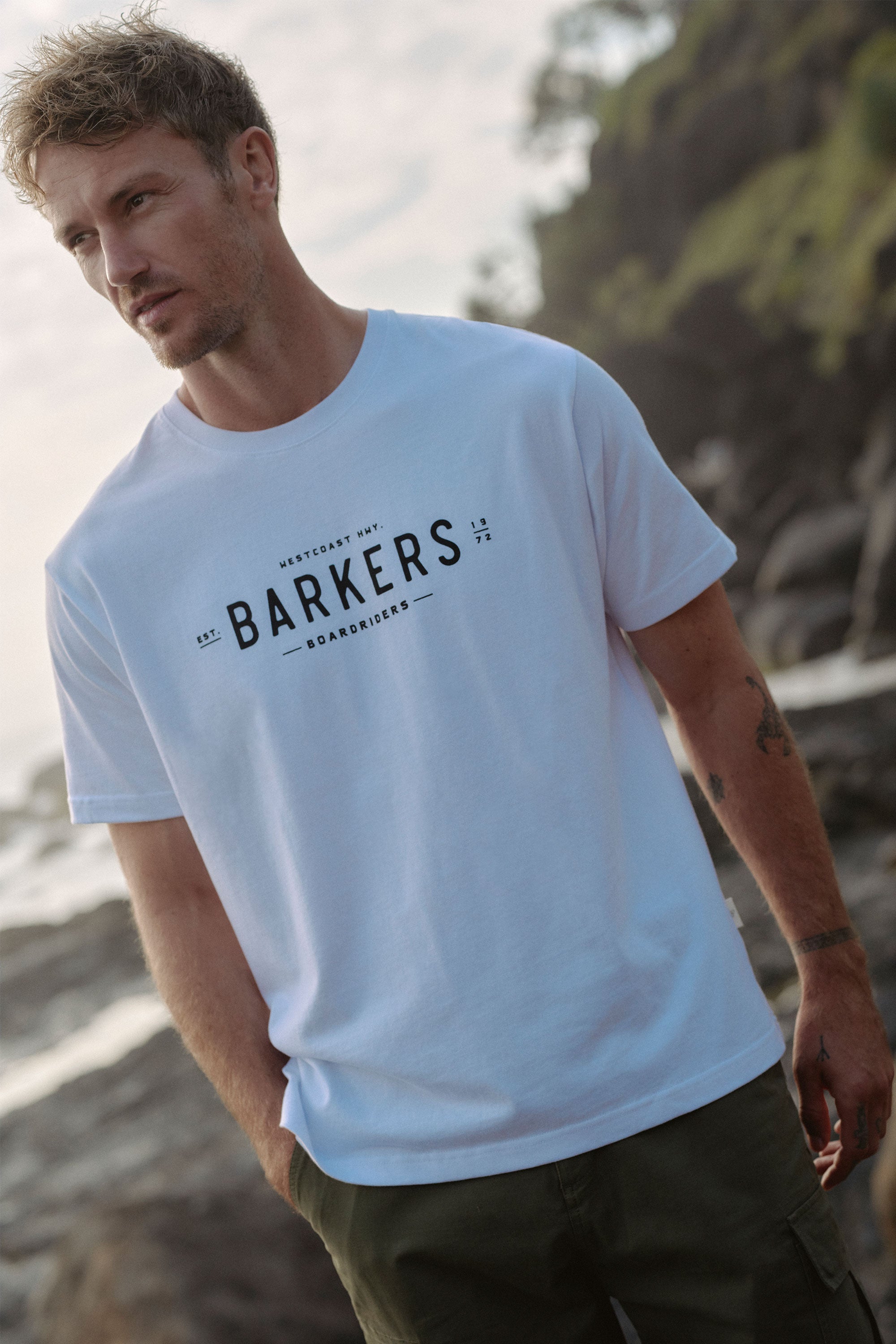 A man with short, light brown hair and stubble stands by a rocky coast, wearing the Barkers Boardriders Logo Tee—an organic cotton t-shirt—and dark olive-green shorts.