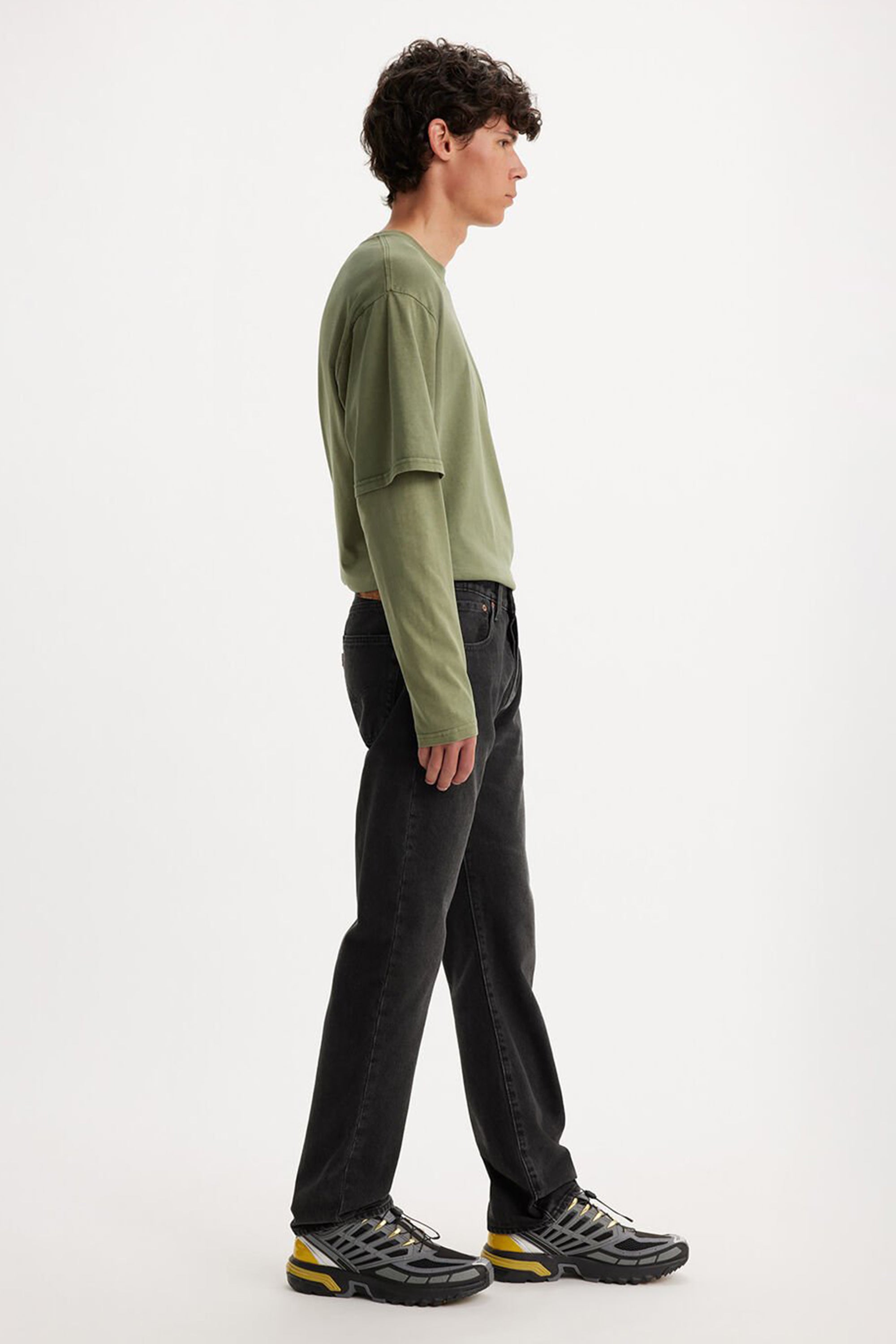 A young man stands in profile against a white background, wearing a loose olive green long-sleeve shirt, Levis 501 Original Jean, and gray running shoes with yellow accents.