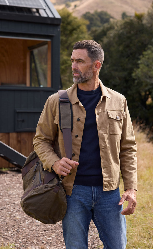 A man with short dark hair and a beard stands outdoors carrying a duffel bag over his shoulder. He wears the Barkers Benson Waxed Cotton Jacket, a navy sweater, and jeans. A small dark cabin and grassy hills appear in the background.