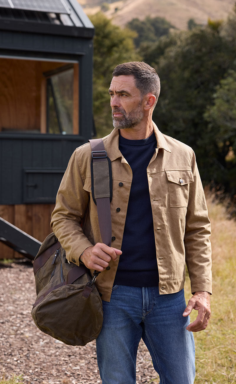 A man with short dark hair and a beard stands outdoors carrying a duffel bag over his shoulder. He wears the Barkers Benson Waxed Cotton Jacket, a navy sweater, and jeans. A small dark cabin and grassy hills appear in the background.