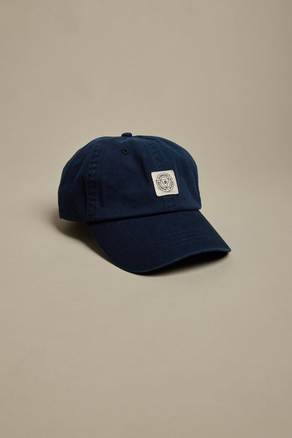 The Barkers Washed Cotton Cap by Barkers is a navy blue vintage wash hat with a curved brim and a small white rectangular patch featuring a circular logo on the front, shown against a plain beige background.