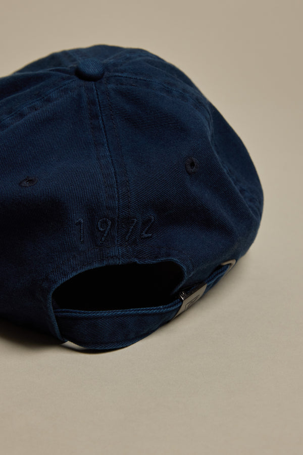 The Barkers Washed Cotton Cap by Barkers is shown in navy blue, viewed from the back with an adjustable strap and "1972" embroidered above the opening, resting on a plain beige surface.