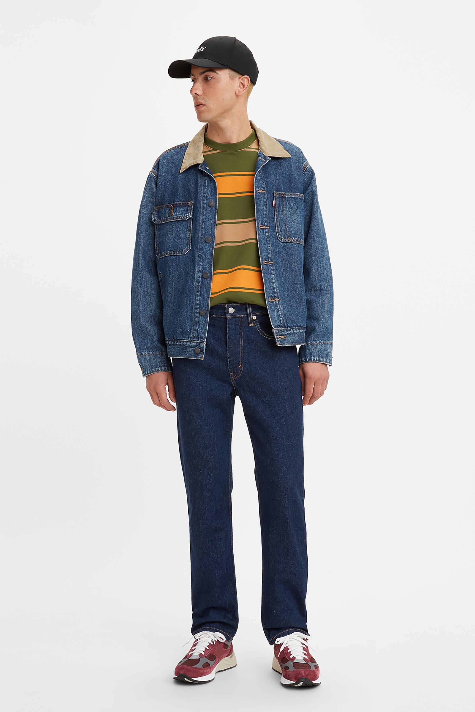 A person stands slightly turned, wearing a black cap, denim jacket with tan collar, striped yellow and green shirt, Levis 516 Straight Jeans, and red sneakers against a white background.