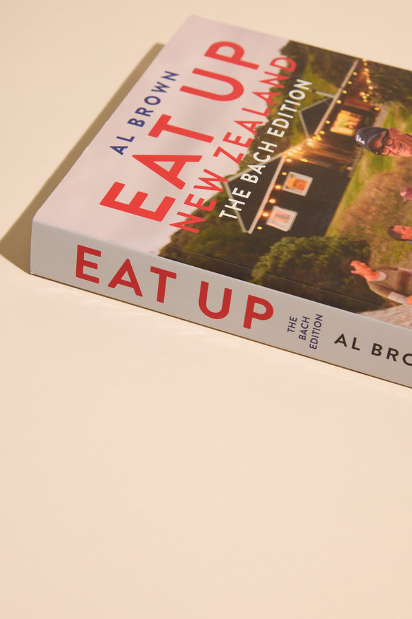 Eat Up New Zealand The Bach Edition by Other rests on a light surface, its cover illustrating a house, greenery, and people—showcasing inviting bach cooking with delicious New Zealand recipes.