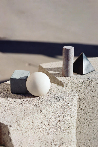 Areaware Drink Rocks by Areaware—a gray cube, white sphere, granite cylinder, and metallic pyramid—rest on light stone surfaces in natural sunlight, evoking elegant cocktail chilling stones.