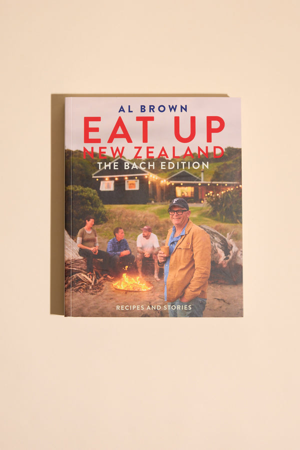 The Other brand’s Eat Up New Zealand The Bach Edition cookbook sits on a beige surface, its cover showing people cooking by a campfire outdoors with a house in the background.