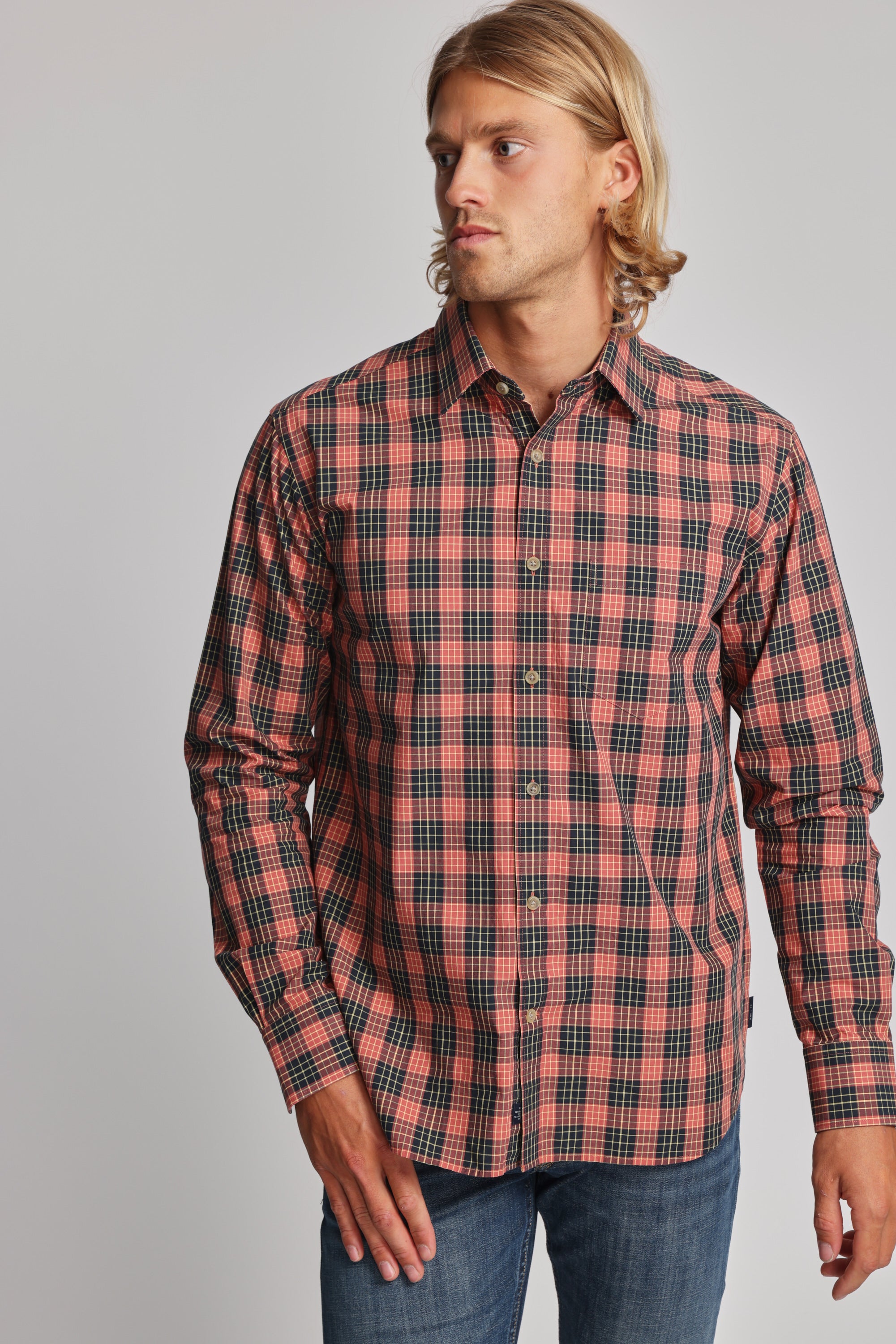 A man with long blonde hair wears the Barkers Formby Long Sleeve Shirt in red and black plaid, paired with blue jeans. He stands against a plain gray background, looking to his left.
