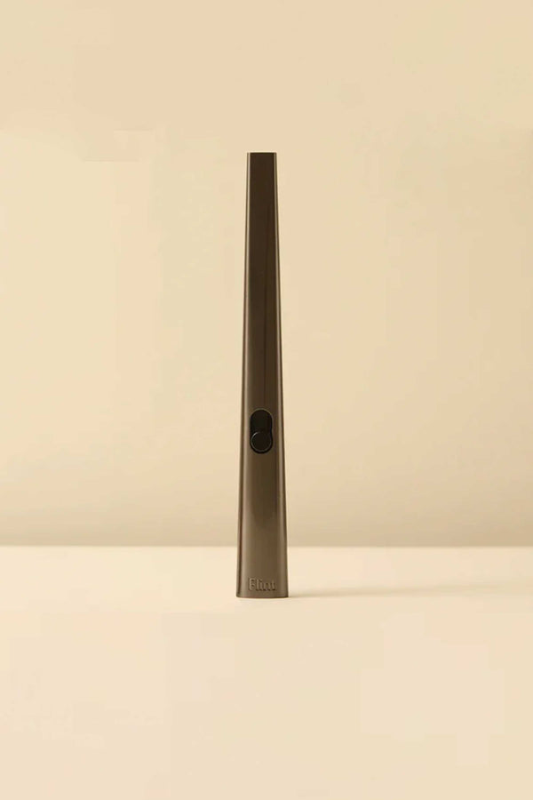 The Flint Rechargeable Lighter 2.0 by Flint is a slim, metallic, wand-shaped gold lighter with a black button, standing upright on a flat beige surface against a matching beige background.