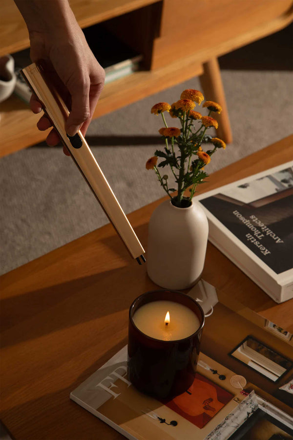 A hand uses the Flint Rechargeable Lighter 2.0 by Flint to light a candle on a wooden coffee table, next to a white vase with yellow flowers and two books.