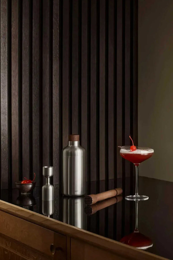 A stylish bar scene featuring the Liquid Lounge Cocktail Jigger by Other, a metal shaker, wooden muddler, bowl of cherries, and a cherry-garnished cocktail atop a glossy black counter framed by dark wood paneling.