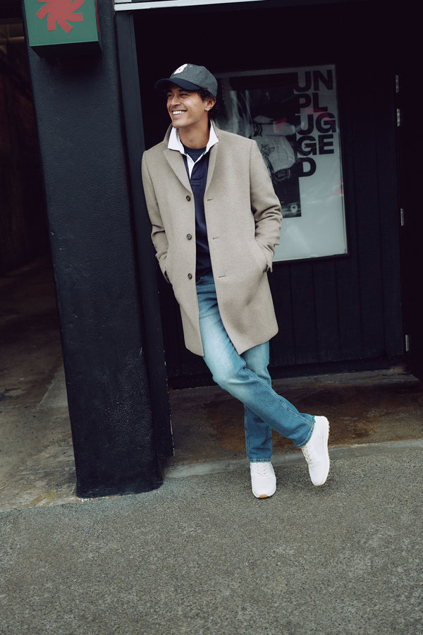 Wearing a Barkers Morrison Coat with blue jeans, white sneakers, and a black cap, a person leans casually against a dark wall by a door, smiling. A poster reading UNPLUGGED is visible behind them.