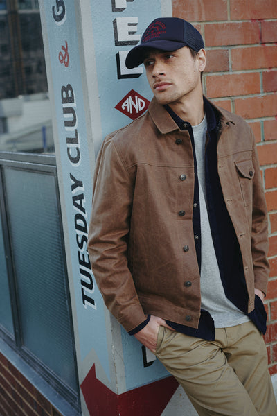 Wearing the Barkers Piston Club Trucker Cap with an embroidered logo, a man leans against a sign and highlights its adjustable fit.