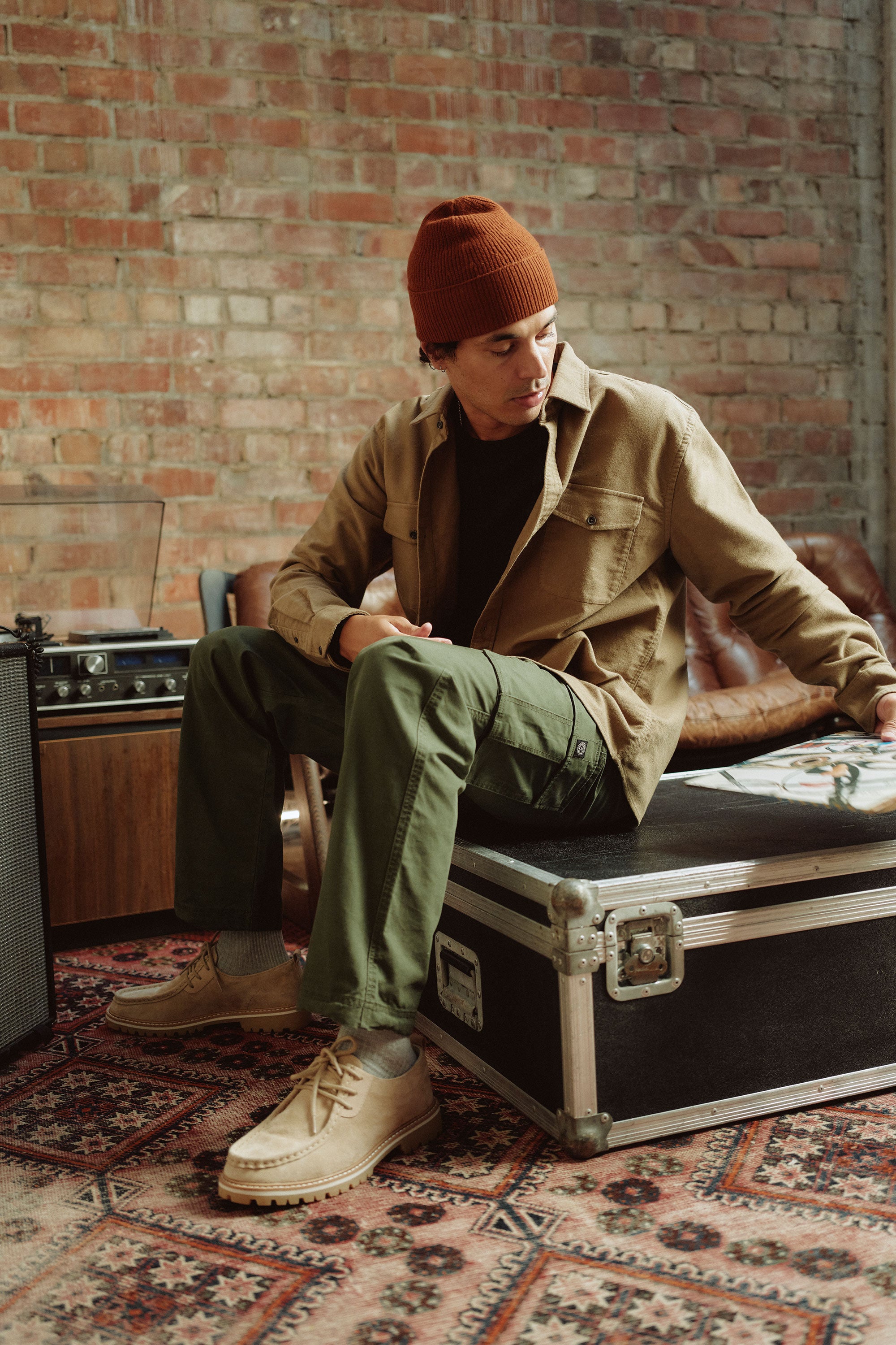 Wearing Barkers Randall Cargo Pants, a person pairs them with a brown beanie, tan cotton jacket, and black shirt while sitting on a black trunk in a cozy room with vintage audio gear and a brick wall, highlighting utilitarian style.