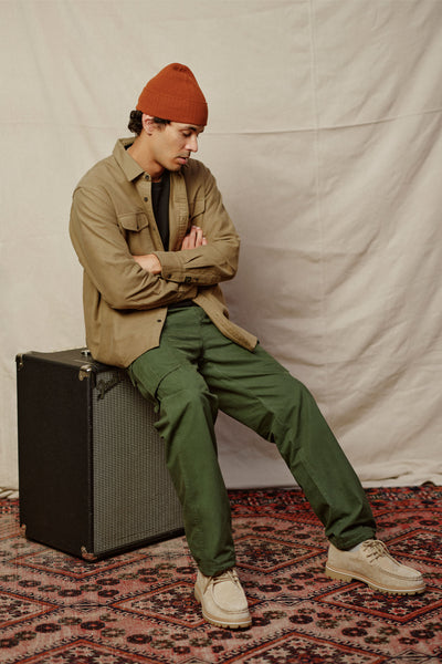 Wearing a rust beanie, tan twill cotton button-up, Barkers Randall Cargo Pants in green, and beige shoes, a person sits cross-armed on a black amplifier against a beige backdrop with patterned rug for a utilitarian look.